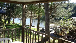 A covered outdoor area with a wooden deck, overlooking a lake with trees and picnic tables. - Camping de Fléchat (Orcival)