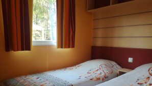 A room with two single beds, a window with curtains, a shelf above the beds, and a wall-mounted light switch. - Camping de Fléchat (Orcival)