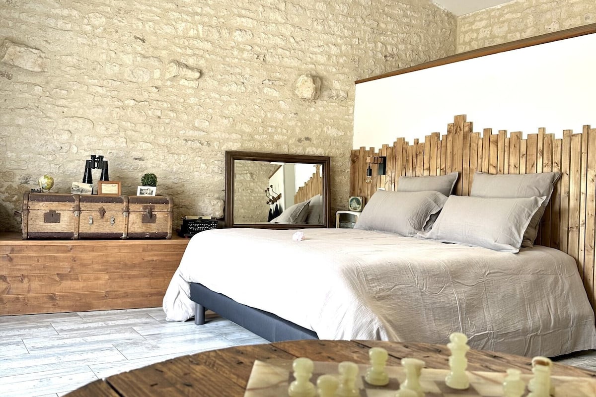 Une chambre avec un mur en pierre, une tête de lit en bois, un lit avec des oreillers, une commode avec des valises, un miroir et un jeu d’échecs.