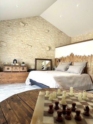 A bedroom with a stone wall, a wooden headboard, a bed with pillows, a dresser with suitcases, a mirror, and a chessboard. - L'Echappee Vendeenne Chambres d'Hotes (Saint-Aubin-la-Plaine)