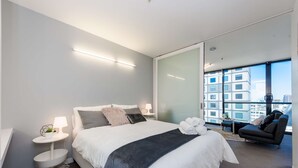 A modern hotel room with a large bed, a seating area with a sofa, a small round table, and a large window offering a city view.
