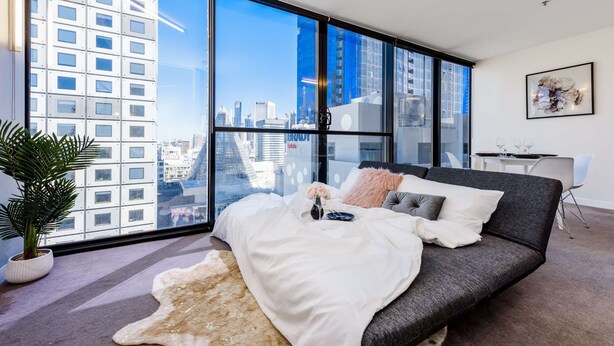 A modern bedroom with a large window, a grey sofa, a white rug, and a potted plant.