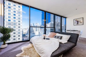 Ein modernes Schlafzimmer mit großem Fenster, einem grauen Sofa, einem weißen Teppich und einer Topfpflanze.