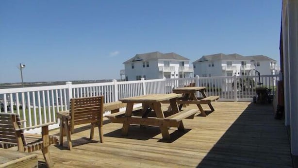 Eine Holzterrasse mit Picknicktischen und Bänken, weißen Geländern und Blick auf weiße Gebäude im Hintergrund.