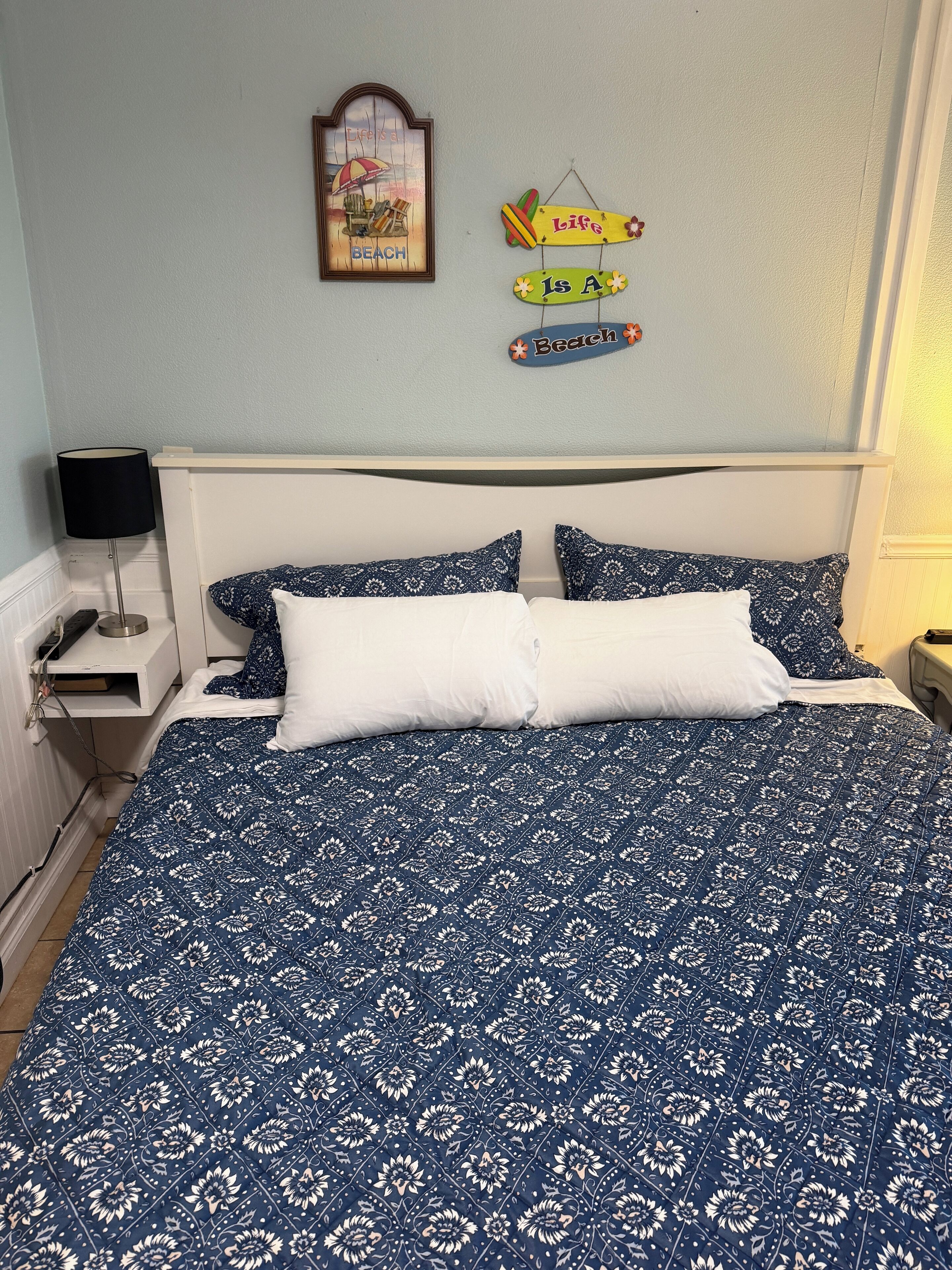 A bedroom with a bed, two pillows, a nightstand, a lamp, and wall decorations including a framed picture and two signs.