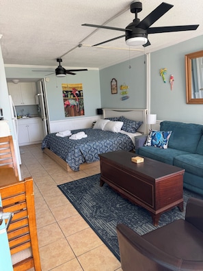 Un espacio habitable compacto con cama, sofá, una silla de madera, una mesita, ventilador de techo, zona de cocina y pared decorada.