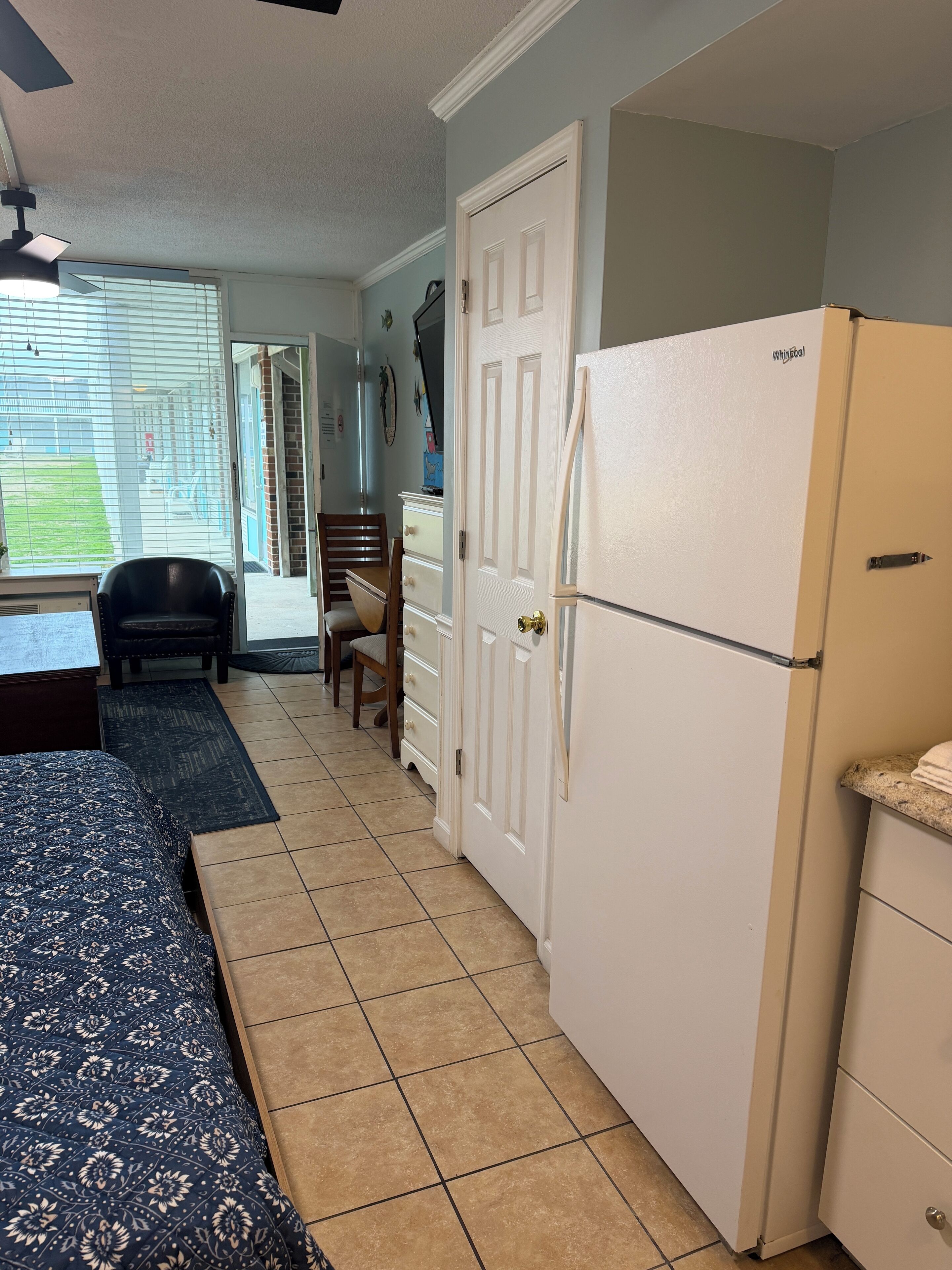 A compact kitchen with a refrigerator, a dining area with a chair and a table, and a window with blinds.