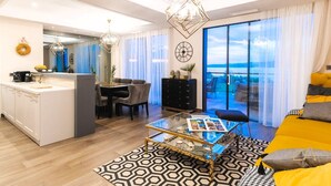 Ein modernes Wohnzimmer mit Glastisch, einer Couch, einem Essbereich mit Stühlen, einer Küche mit weißer Arbeitsplatte und einem großen Fenster mit Blick.