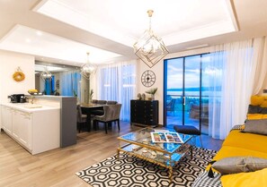 A modern living room with a glass coffee table, a sofa, a dining area with chairs, a kitchen with a white countertop, and a large window with a view.
