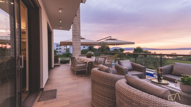 Ein Balkon mit Korbmöbeln, einem Tisch mit Flasche und Gläsern sowie Blick auf das Meer bei Sonnenuntergang.