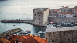 Una fortaleza de piedra histórica con un foso y una bandera, con vistas a un puerto con barcos y un muelle.