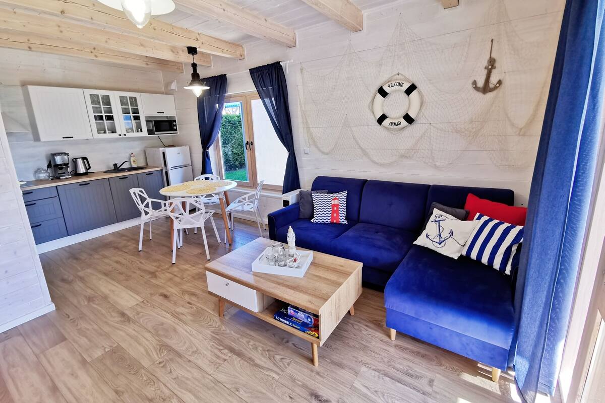 Een gezellige woonkamer met een blauwe bank, een houten salontafel en een witte eettafel met stoelen. Er is een keuken met kastjes en een magnetron.