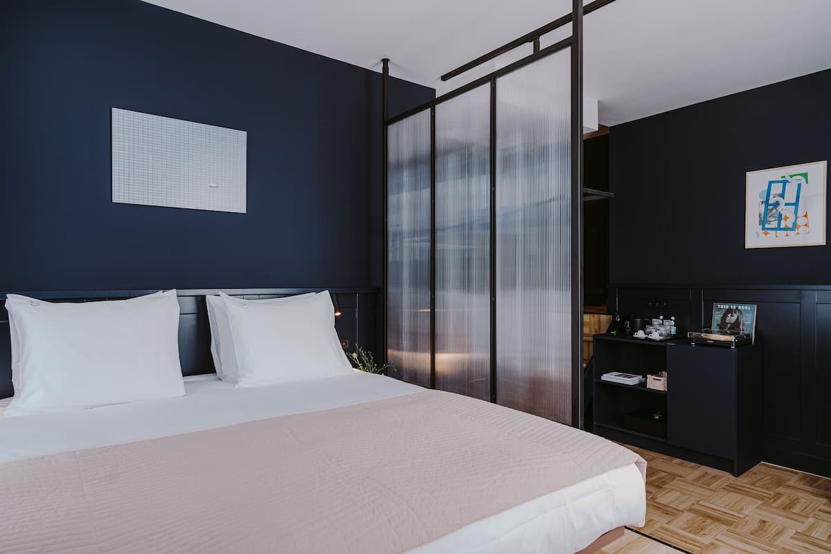 Ein modernes Schlafzimmer mit einem großen Bett, einer dunklen Wand, einem Fenster mit Jalousien und einem kleinen Regal mit Gegenständen.