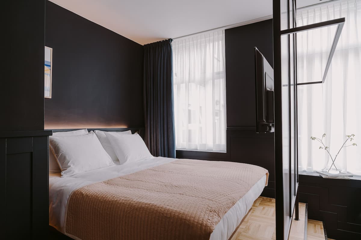 Ein modernes Hotelzimmer mit einem großen Bett, einem Fenster mit leichtem Vorhang und Blick auf die Stadt.