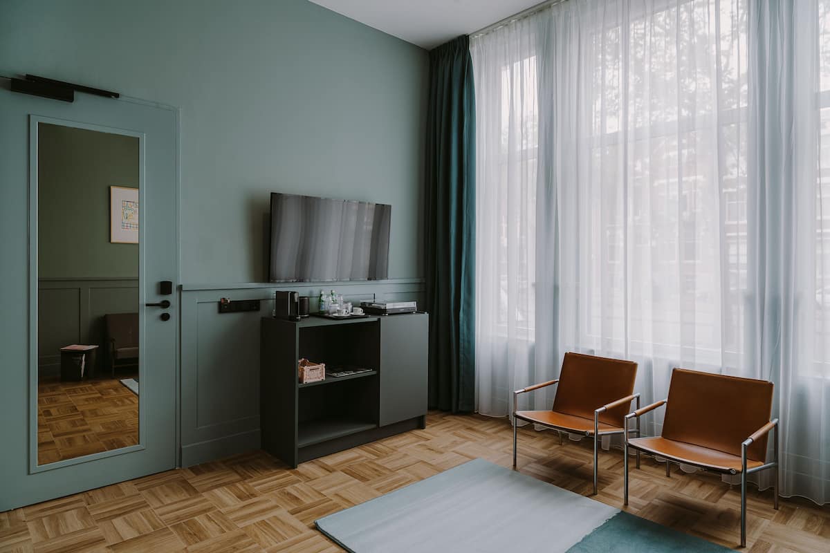 Ein Zimmer mit Holzboden im Fischgrätmuster, einem grauen Sofa, zwei braunen Ledersesseln, einem kleinen Tisch, einem Fernseher auf einem Schrank und einem Fenster mit durchscheinenden Vorhängen.