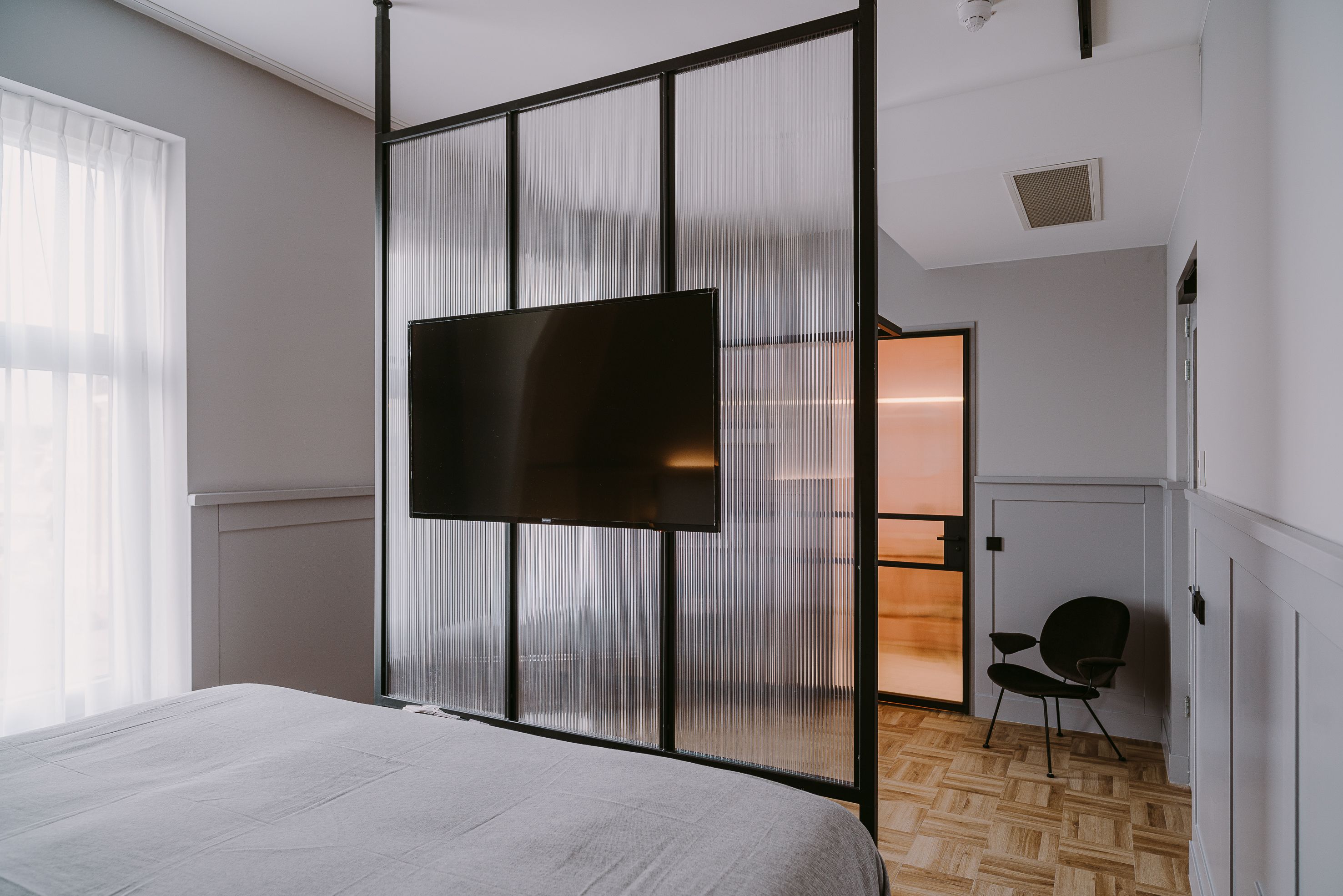 A modern hotel room with a large bed, a TV mounted on a sliding screen wall, a chair, and a wooden herringbone floor.