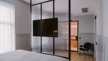 A modern hotel room with a large bed, a TV mounted on a sliding screen wall, a chair, and a wooden herringbone floor.