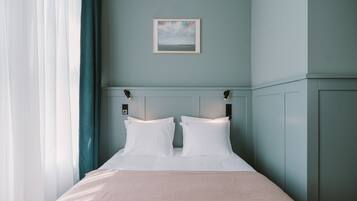 A neatly made bed with white linens and two pillows, a framed picture on the wall, and a window with sheer curtains.