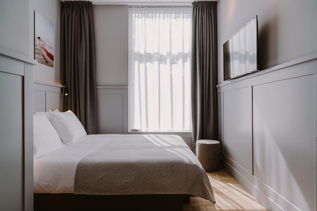 Ein ordentlich eingerichtetes Schlafzimmer mit Bett, Nachttisch, einem Fenster mit Vorhängen und einem Wand-Fernseher.