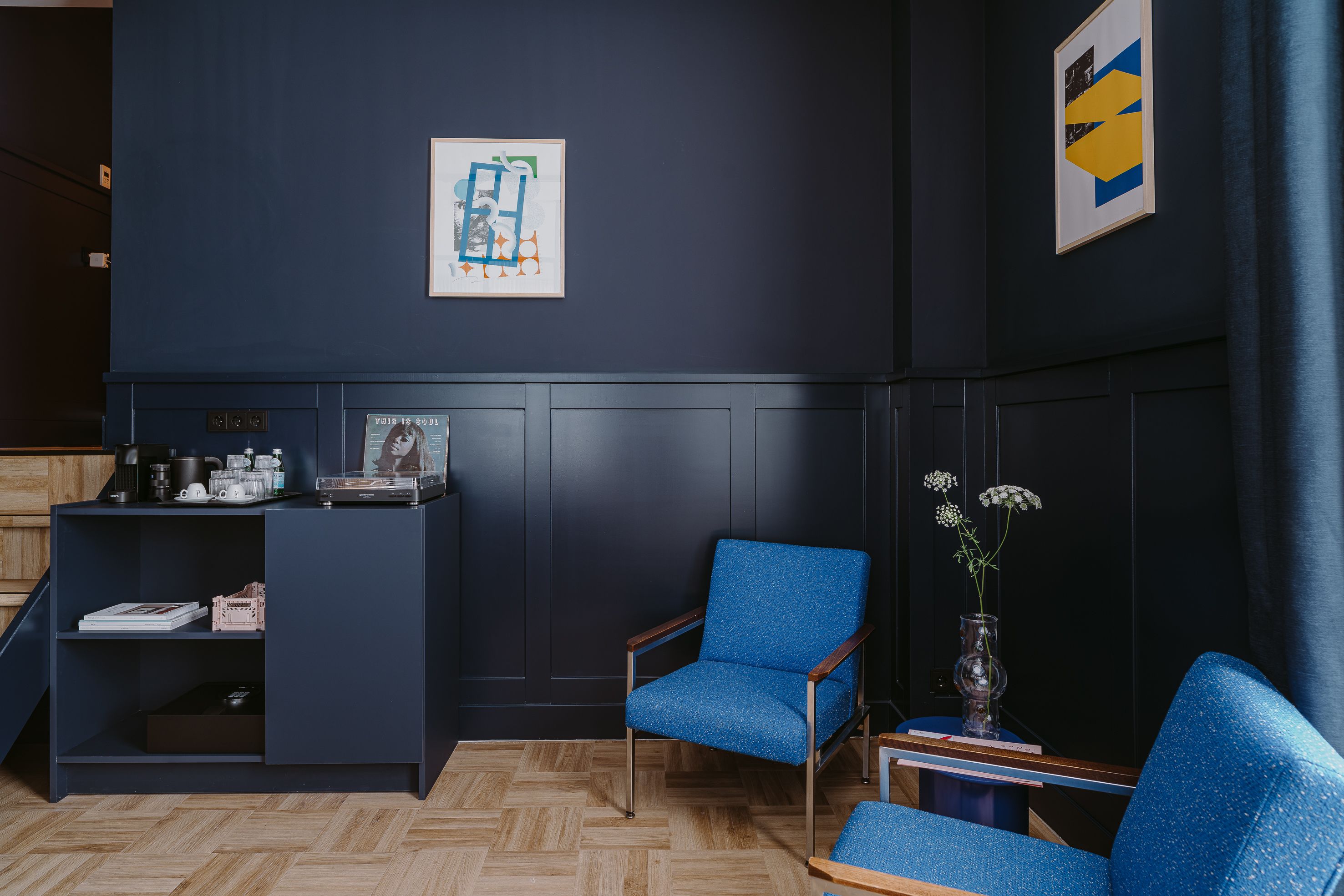 Quarto com decoração escura, uma poltrona estofada azul, uma mesa pequena com um vaso e um armário de madeira com diversos objetos.