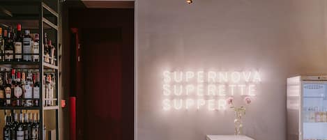 Un coin bar agrémenté d’un panneau néon sur lequel on peut lire « SUPERNOVA SUPERETTE SUPPER », d’un présentoir à bouteilles et d’un réfrigérateur rempli de bouteilles.