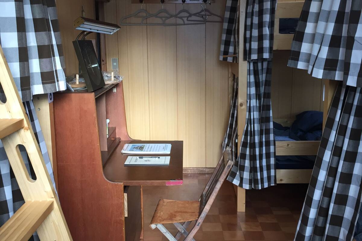 A compact room with a bunk bed, a desk with a chair, a small table, and a hanging light fixture.
