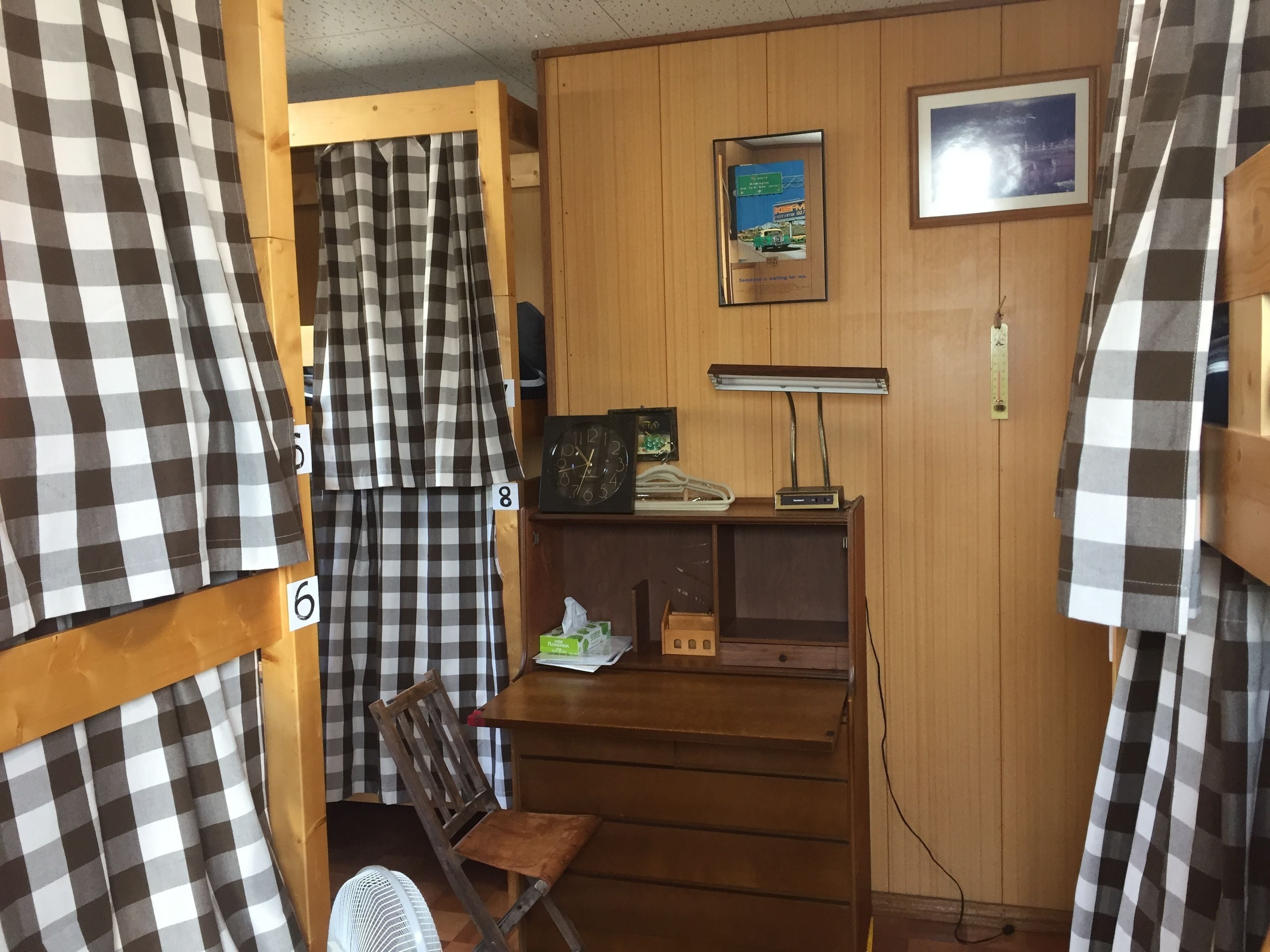 A small, compact room with a wooden desk, a chair, a clock, a framed picture, and a fan.