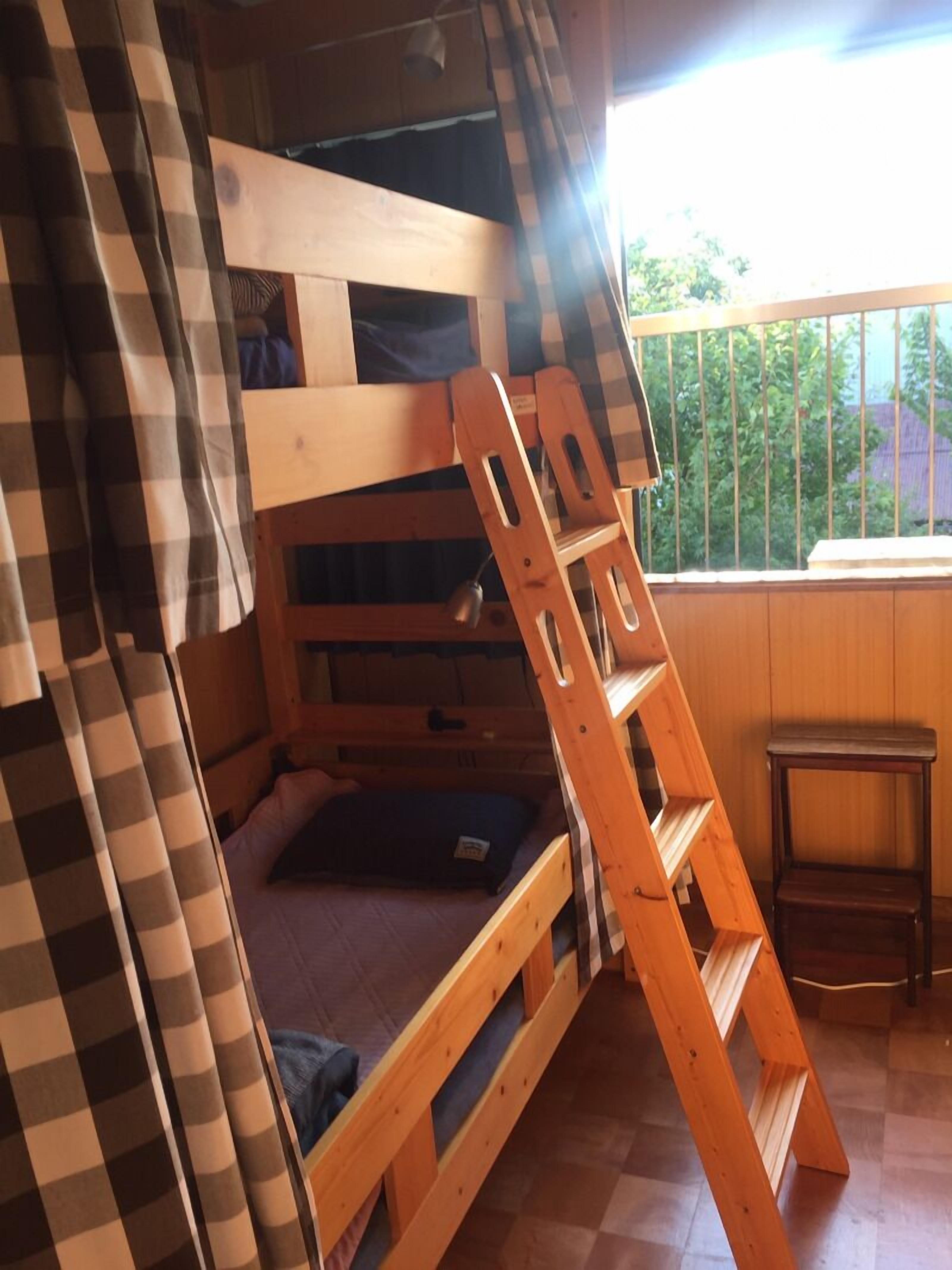 Kerrossänky, jossa on puinen tikapuu, ruutukuviollinen verho ja ikkuna, josta avautuu näkymä vehreään maisemaan.