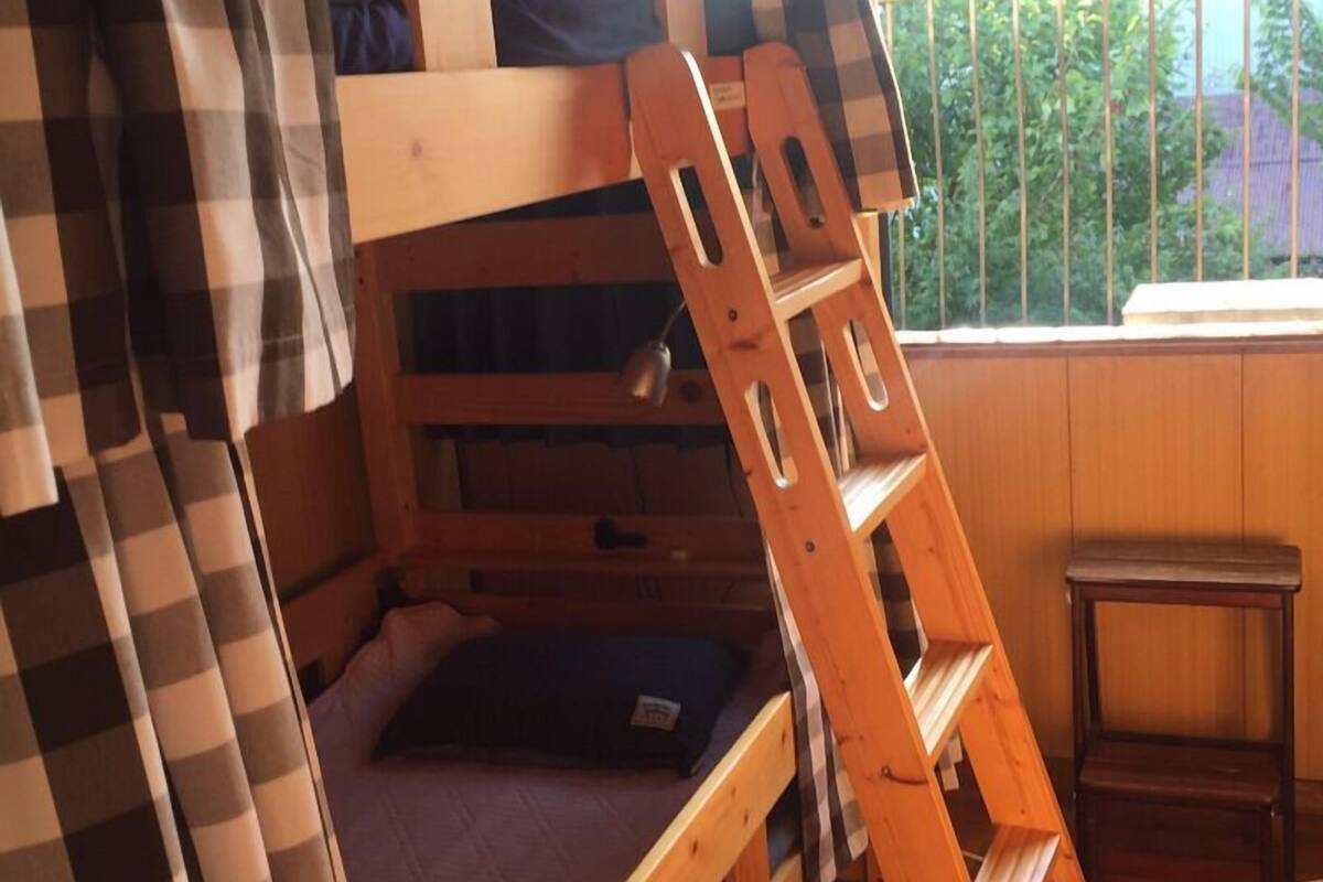 A bunk bed with a wooden ladder, a checkered curtain, and a window with a view of greenery.