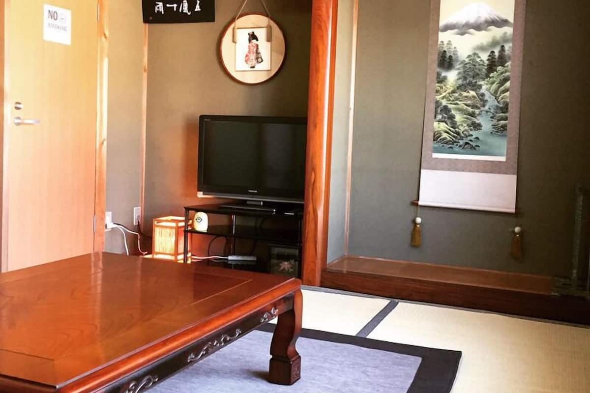 Une pièce de style japonais traditionnel, avec un sol recouvert de tatamis, un téléviseur, un tableau encadré au mur et une vue sur une montagne par la fenêtre.