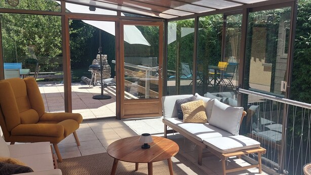 Un patio soleado con un sofá, un sillón y una mesa redonda, con vistas a un jardín trasero que tiene mobiliario exterior y una parrilla.