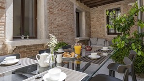 Un espace extérieur aménagé pour les repas, avec un mur en briques, une table en verre et des chaises. On peut voir une carafe, des verres et un saladier rempli de fruits sur la table.