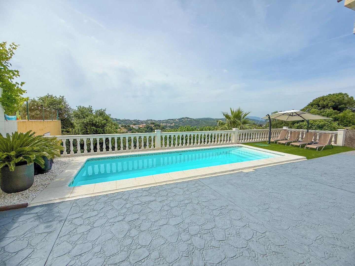 Une piscine rectangulaire dotée d’une balustrade blanche, entourée d’une terrasse au revêtement imitant la pierre, et offrant une vue sur des collines lointaines et la verdure.