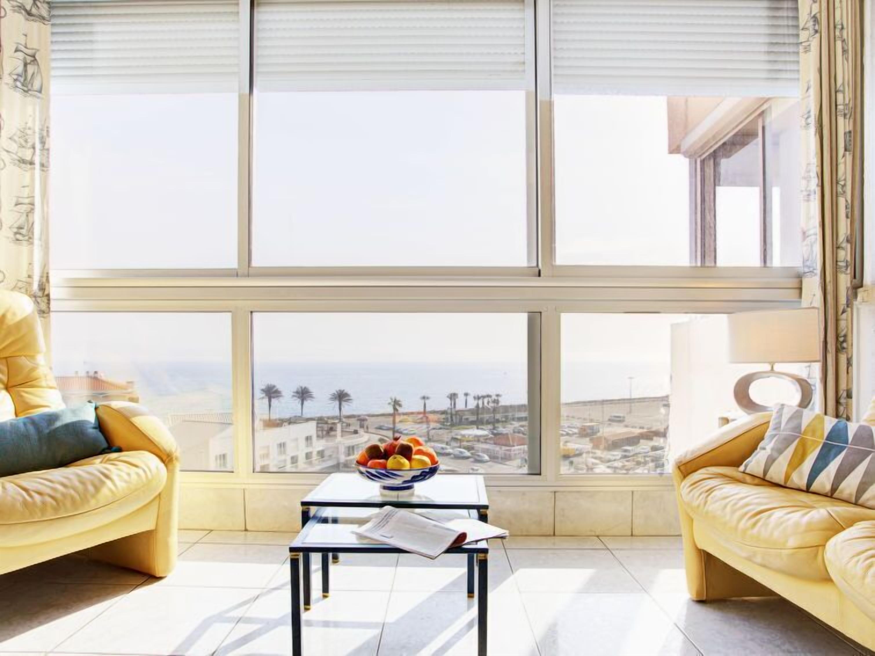 Ein modernes Wohnzimmer mit einem großen Fenster, zwei gelben Ledersofas, einem kleinen Tisch mit Obstschale und einer Lampe.