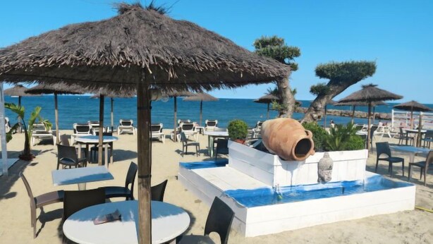 Un restaurant situé en bord de mer, doté de parasols en chaume, de tables en plein air et d’un élément décoratif aquatique.