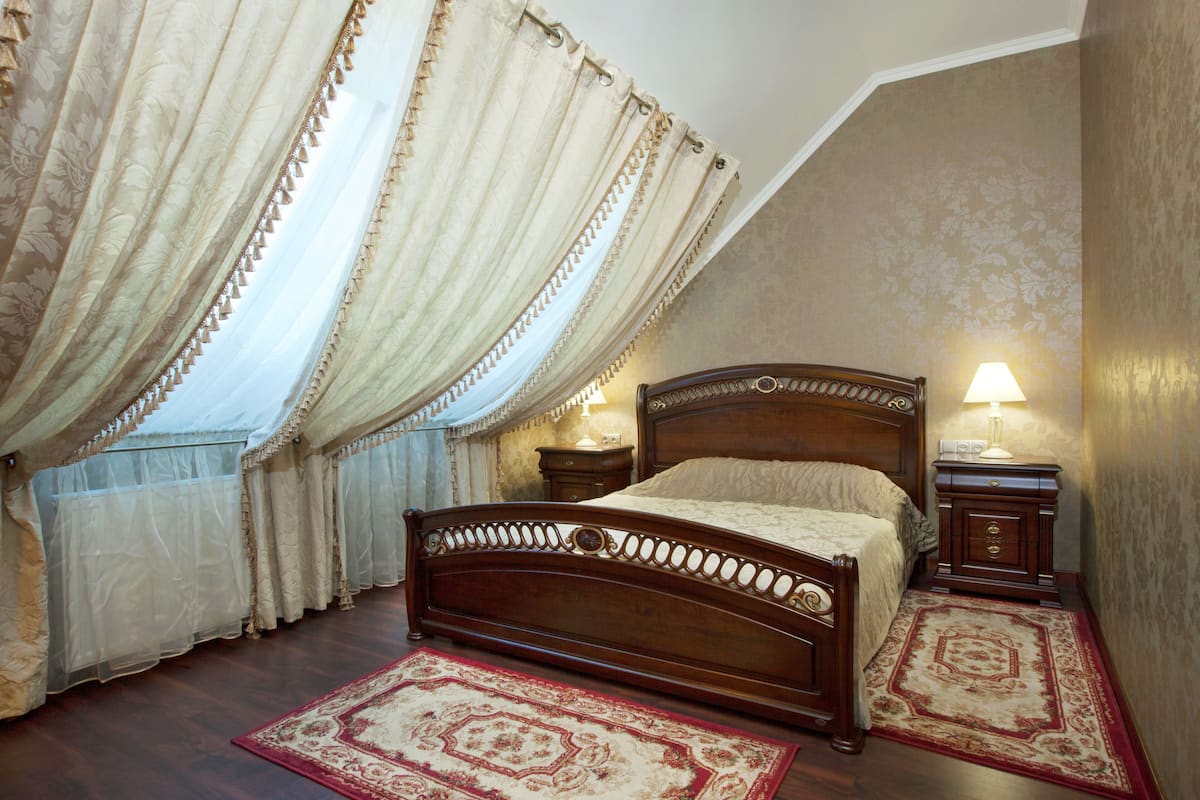Une chambre à coucher avec un lit en bois, deux tables de chevet, une commode, une fenêtre avec des rideaux légers et un tapis à motifs.