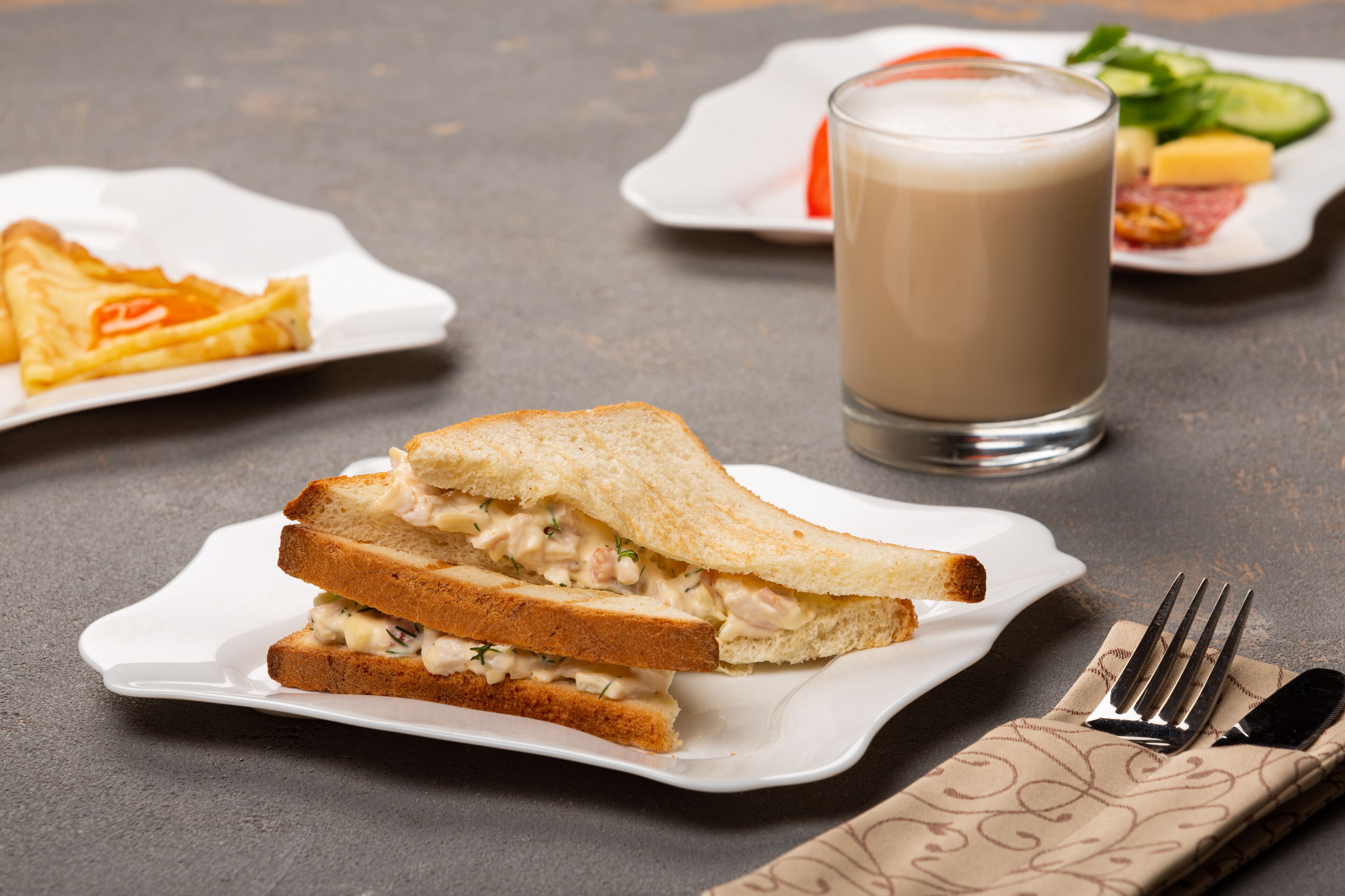 A sandwich on a white plate, a glass of milk, and a fork on a napkin.