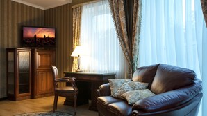 A living room with a leather sofa, a wooden cabinet, a desk, and a television displaying a cityscape at sunset. - Hotel Helicopter (Kremenchuk)