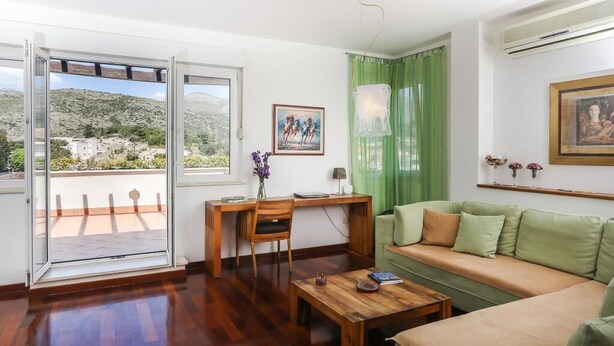 A living room with a wooden desk, a green sofa, a wooden coffee table, and a view of a mountainous landscape through a glass door. - Apartment Alma - Two Bedroom Apartment with Terrace and Sea View (Dubrovnik)