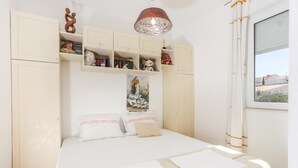 A bedroom with a bed, white curtains, a window, and a shelf with various items. - Apartment Alma - Two Bedroom Apartment with Terrace and Sea View (Dubrovnik)