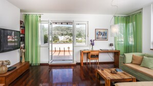 A living room with a wooden floor, a sofa, a coffee table, a TV, a desk with a chair, a bookshelf, and a view through a glass door to an outdoor balcony. - Apartment Alma - Two Bedroom Apartment with Terrace and Sea View (Dubrovnik)