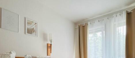 Habitación de hotel con cama, escritorio y silla. Cortinas en la ventana y una lámpara de pared.