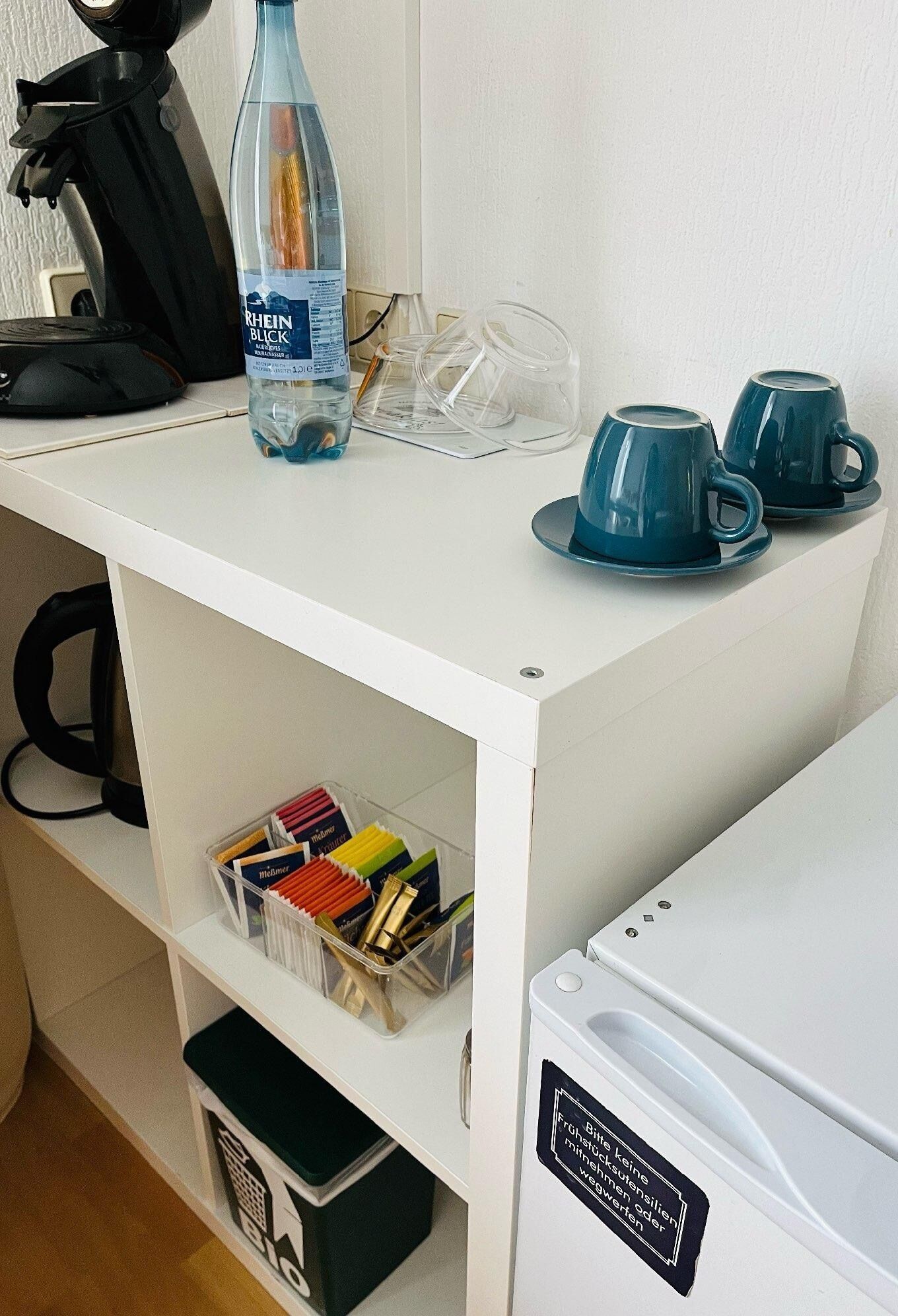 Una kitchenette con una encimera blanca, una botella de agua, una cafetera y una máquina de café.