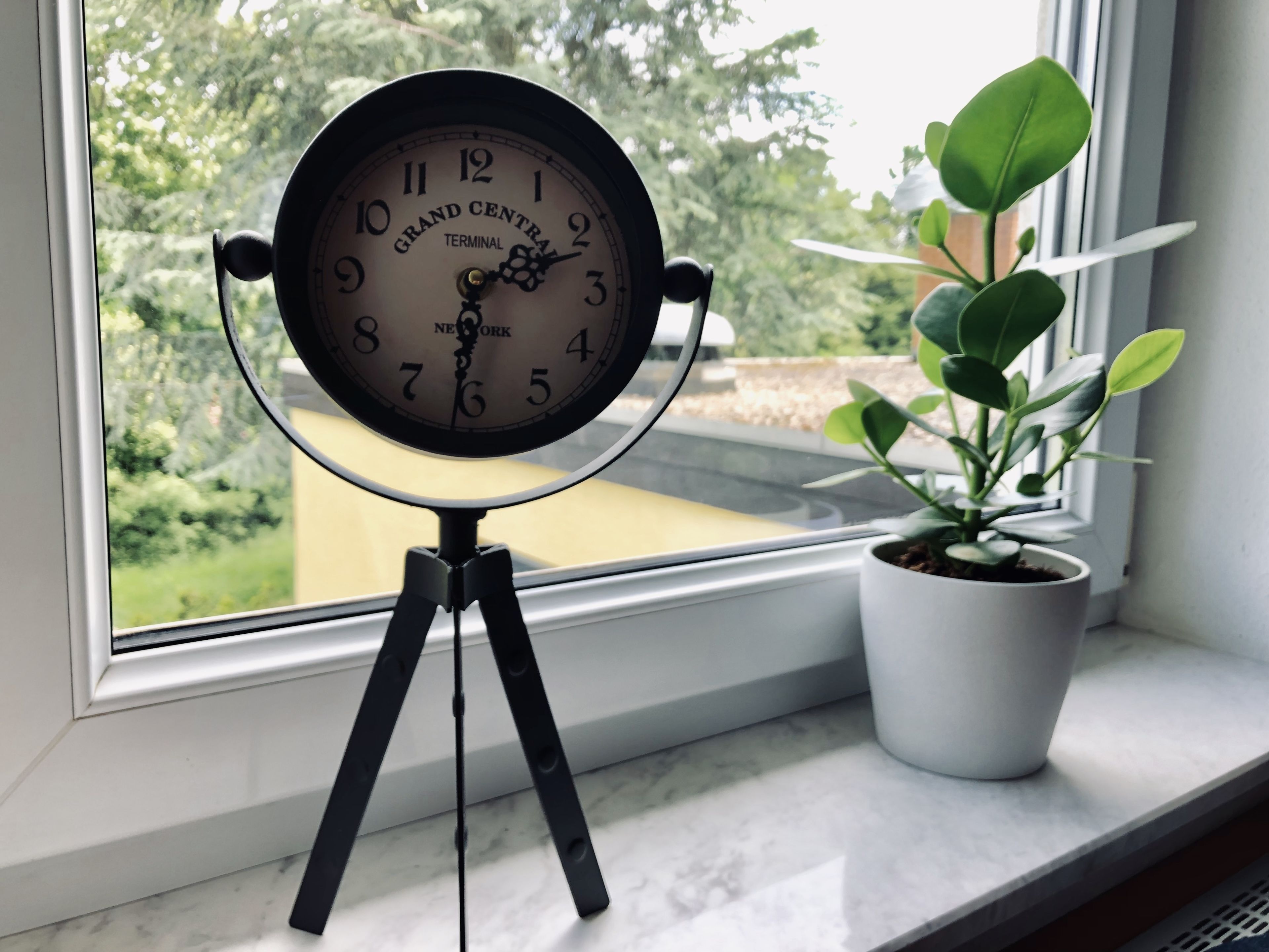Un reloj de Grand Central Terminal sobre un trípode, junto a una ventana con una planta en maceta.