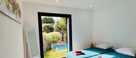 Ein modernes Schlafzimmer mit einem großen Bett, einer kleinen Sitzecke und Blick auf einen Garten durch eine Glastür.