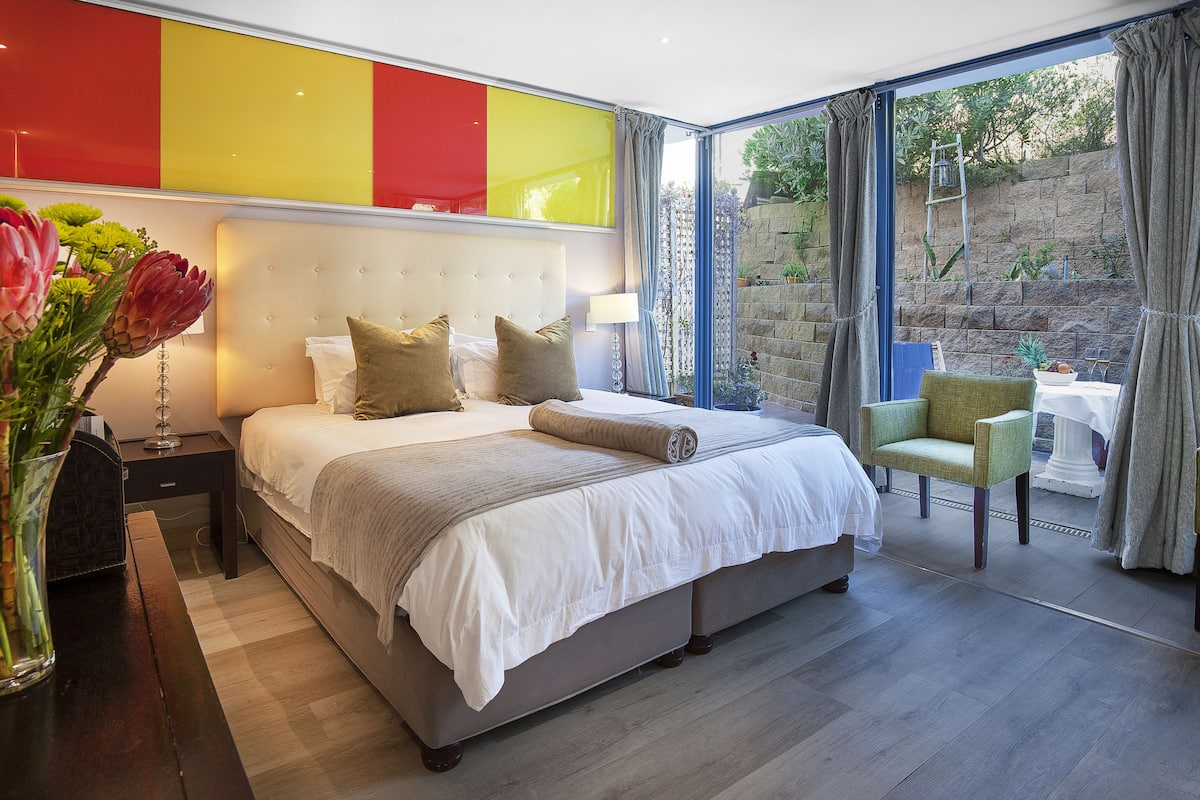 Un dormitorio moderno con una cama grande, mesitas de noche, un jarrón de flores y vistas a un jardín a través de amplias ventanas.