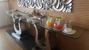 A buffet setup with various food items and beverages on a glass table against a wooden wall with a decorative mirror above. - Magellans Hotel (Cape Town)