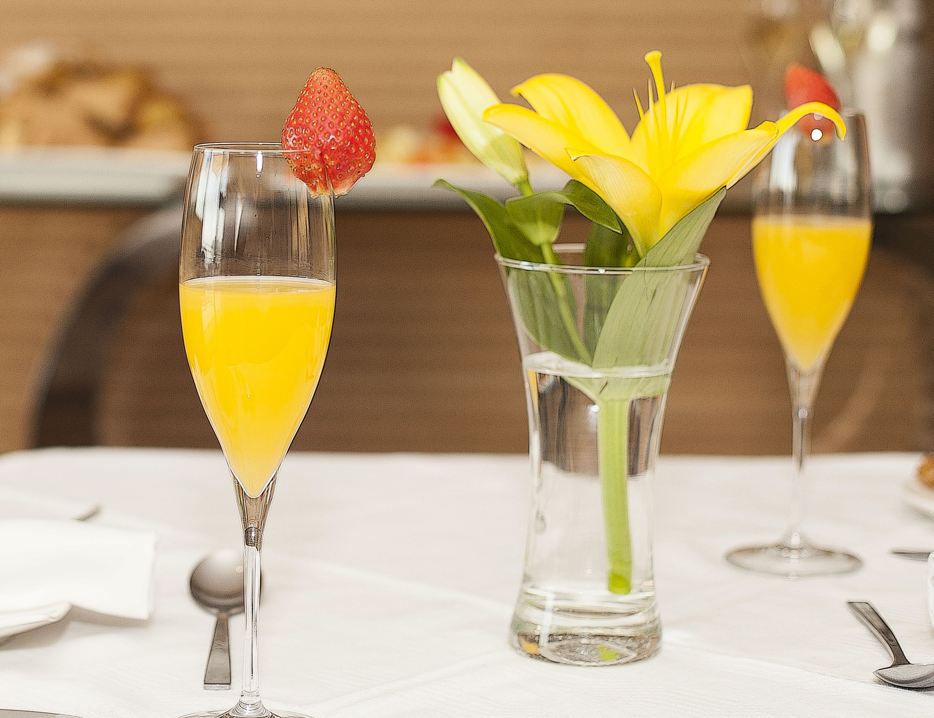Dos vasos de jugo de naranja, uno con una fresa como adorno, y un jarrón con flores amarillas.