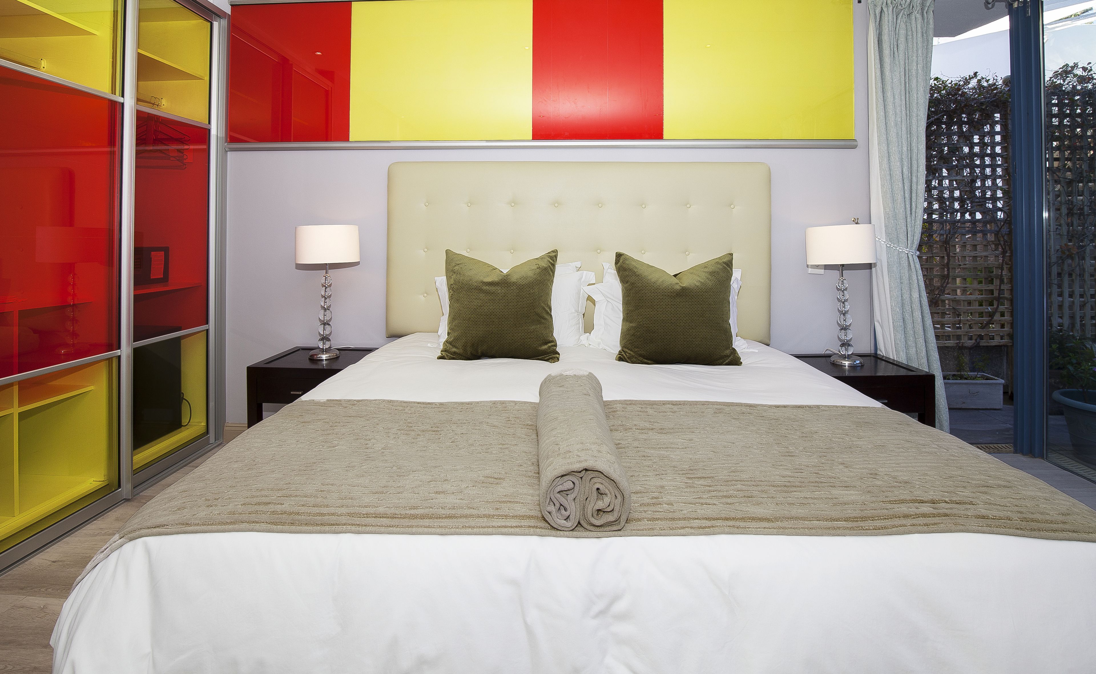 Quarto de hotel moderno com uma cama grande, duas mesas de cabeceira e uma parede em vermelho e amarelo vibrantes.
