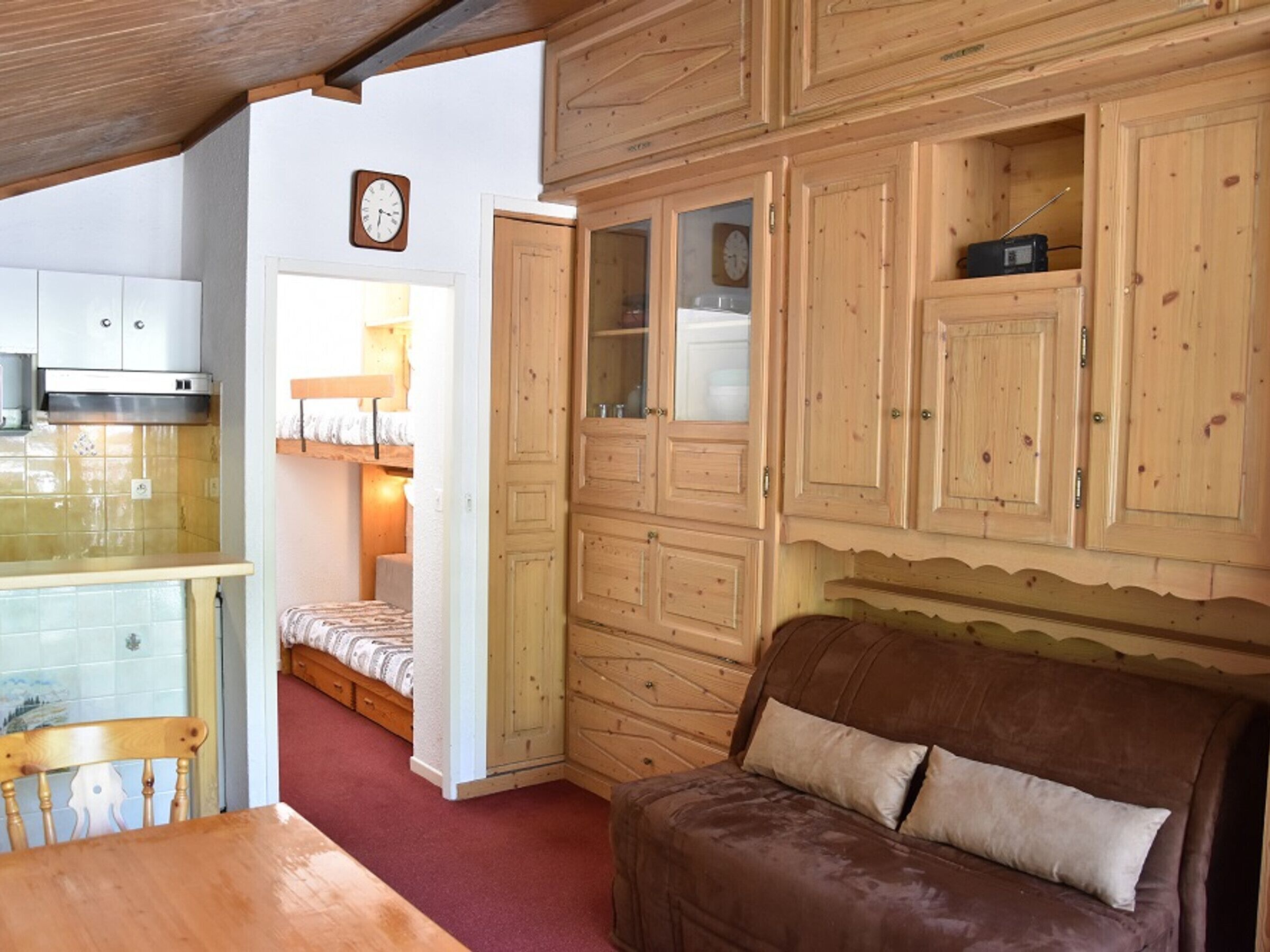 Ein gemütliches Zimmer mit einer hölzernen Bank, einem braunen Sofa und einer Küchenzeile mit Schränken und einer Uhr.